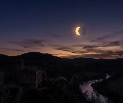 Twilight over a stone village and winding river valley as a solar eclipse forms a bright crescent in the sky behind distant hills.