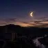 Twilight over a stone village and winding river valley as a solar eclipse forms a bright crescent in the sky behind distant hills.