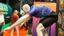 Mannequin in a blue athletic jersey bent forward on a trade show display with people and orange signage in the background.
