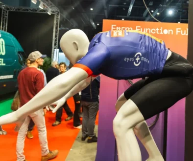 Mannequin in a blue athletic jersey bent forward on a trade show display with people and orange signage in the background.