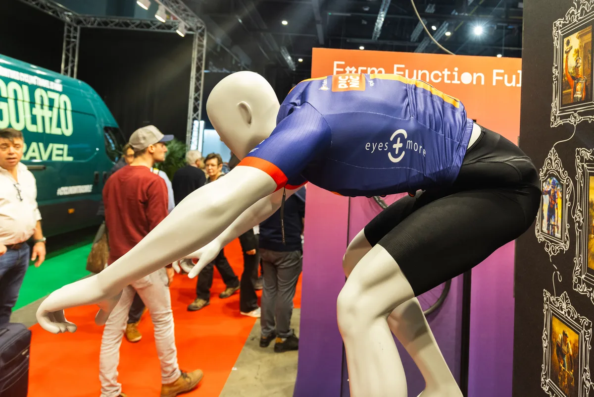 Mannequin in a blue athletic jersey bent forward on a trade show display with people and orange signage in the background.