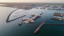 Aerial view of a large port with breakwaters, docks, and cranes under construction along calm harbor waters at sunset.