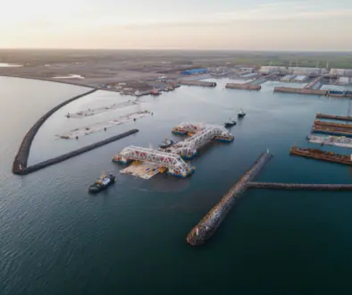 Aerial view of a large port with breakwaters, docks, and cranes under construction along calm harbor waters at sunset.