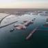 Aerial view of a large port with breakwaters, docks, and cranes under construction along calm harbor waters at sunset.
