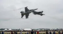 Two gray fighter jets perform a close, mid-air maneuver above a crowded airshow with white tents and spectators below.