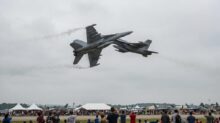 Two gray fighter jets perform a close, mid-air maneuver above a crowded airshow with white tents and spectators below.