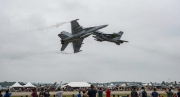 Two gray fighter jets perform a close, mid-air maneuver above a crowded airshow with white tents and spectators below.