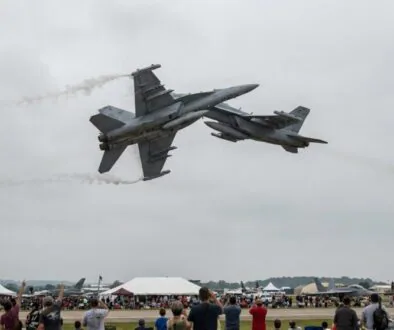 Two gray fighter jets perform a close, mid-air maneuver above a crowded airshow with white tents and spectators below.