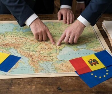 Two people in business suits point at a detailed Europe map on a wooden table, with Romanian and Moldovan flags nearby.