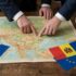Two people in business suits point at a detailed Europe map on a wooden table, with Romanian and Moldovan flags nearby.