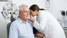 Eye care professional adjusts a phoropter while an older man sits in an exam chair during a vision test, with a chart visible in the background.