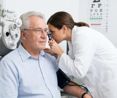 Eye care professional adjusts a phoropter while an older man sits in an exam chair during a vision test, with a chart visible in the background.