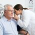 Eye care professional adjusts a phoropter while an older man sits in an exam chair during a vision test, with a chart visible in the background.