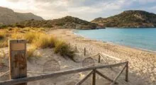 Sunny beach with a wooden rope fence, sand dunes, and tufts of grass leading to clear turquoise water and rocky hills in the distance.