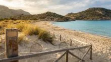 Sunny beach with a wooden rope fence, sand dunes, and tufts of grass leading to clear turquoise water and rocky hills in the distance.