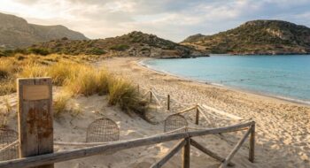 Sunny beach with a wooden rope fence, sand dunes, and tufts of grass leading to clear turquoise water and rocky hills in the distance.