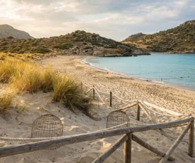 Sunny beach with a wooden rope fence, sand dunes, and tufts of grass leading to clear turquoise water and rocky hills in the distance.