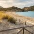 Sunny beach with a wooden rope fence, sand dunes, and tufts of grass leading to clear turquoise water and rocky hills in the distance.
