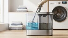 Self-wringing mop bucket with a blue-water reservoir and mop attached, set on a wooden floor beside a front-loading washing machine in a laundry room.