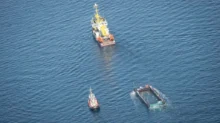 Aerial view of a yellow-and-white work ship with a small tug nearby and a rectangular barge partially submerged in deep blue ocean.