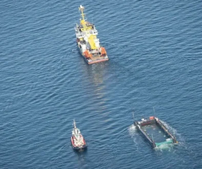 Aerial view of a yellow-and-white work ship with a small tug nearby and a rectangular barge partially submerged in deep blue ocean.