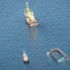 Aerial view of a yellow-and-white work ship with a small tug nearby and a rectangular barge partially submerged in deep blue ocean.