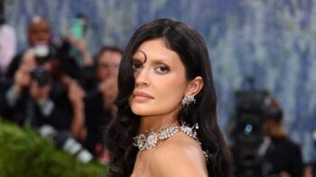 Elegant woman on the red carpet wearing an elaborate pearl necklace and earrings, photographers in the background.