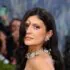 Elegant woman on the red carpet wearing an elaborate pearl necklace and earrings, photographers in the background.