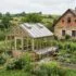 Garden scene with a wooden-framed greenhouse beside a stone cottage; a person tends raised veggie beds amid flowers.