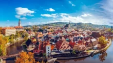 Panoramic view of a riverside historic town with red-tiled roofs, church spires, and autumn trees against a bright blue sky.