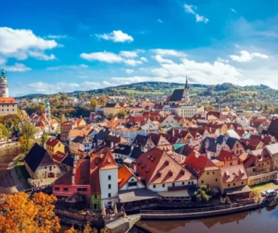 Panoramic view of a riverside historic town with red-tiled roofs, church spires, and autumn trees against a bright blue sky.