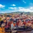 Panoramic view of a riverside historic town with red-tiled roofs, church spires, and autumn trees against a bright blue sky.