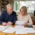 Couple reviewing several documents at a wooden dining table in a living room, using a calculator nearby.