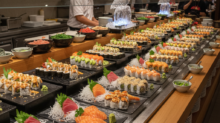 Row of sushi platters with assorted nigiri and rolls lined along a long buffet counter, chefs visible in the background.