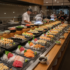 Row of sushi platters with assorted nigiri and rolls lined along a long buffet counter, chefs visible in the background.