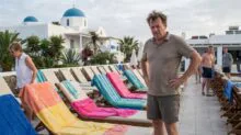 Man with hands on hips stands beside a row of colorful beach chairs on a pool deck at a white-blue domed resort.