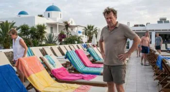 Man with hands on hips stands beside a row of colorful beach chairs on a pool deck at a white-blue domed resort.