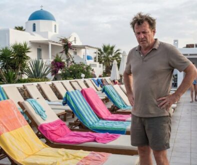 Man with hands on hips stands beside a row of colorful beach chairs on a pool deck at a white-blue domed resort.