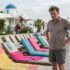 Man with hands on hips stands beside a row of colorful beach chairs on a pool deck at a white-blue domed resort.