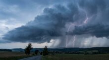 Dark storm clouds over a rural landscape with a winding road; bright lightning bolts strike near the horizon.