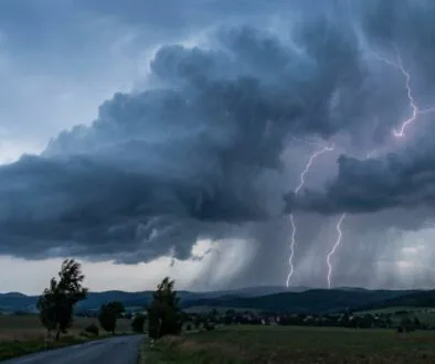 Dark storm clouds over a rural landscape with a winding road; bright lightning bolts strike near the horizon.