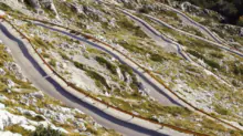 Winding mountain road with sharp switchbacks and wooden guardrails along rocky, grassy slopes.