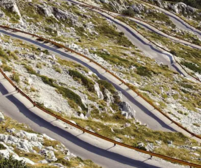 Winding mountain road with sharp switchbacks and wooden guardrails along rocky, grassy slopes.