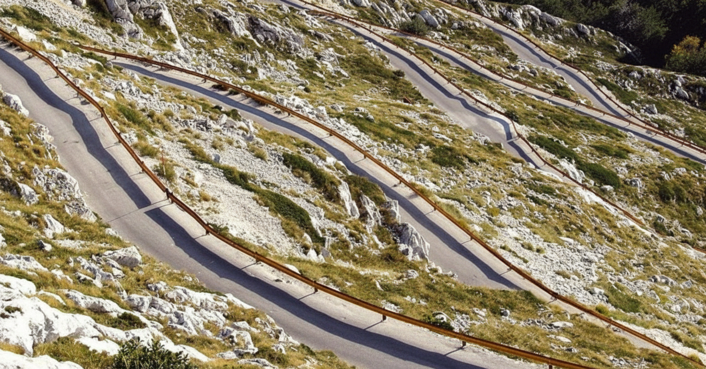 Winding mountain road with sharp switchbacks and wooden guardrails along rocky, grassy slopes.
