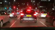 Nighttime city traffic from a car’s dashboard view; red brake lights and cars ahead on a multi-lane road.