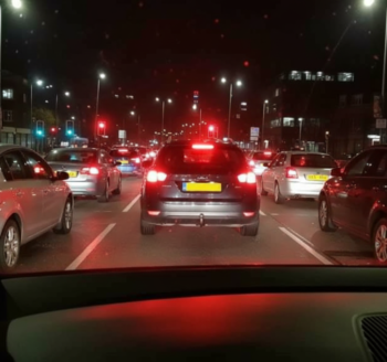 Nighttime city traffic from a car’s dashboard view; red brake lights and cars ahead on a multi-lane road.
