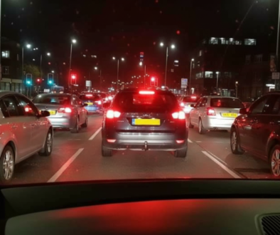 Nighttime city traffic from a car’s dashboard view; red brake lights and cars ahead on a multi-lane road.