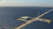 A vast solar farm with rows of blue photovoltaic panels, a central white building, dirt roads, and power transmission towers."The horizon shows more panels stretching to the distance.