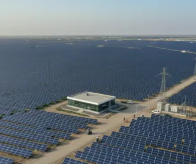 A vast solar farm with rows of blue photovoltaic panels, a central white building, dirt roads, and power transmission towers."The horizon shows more panels stretching to the distance.