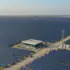A vast solar farm with rows of blue photovoltaic panels, a central white building, dirt roads, and power transmission towers."The horizon shows more panels stretching to the distance.